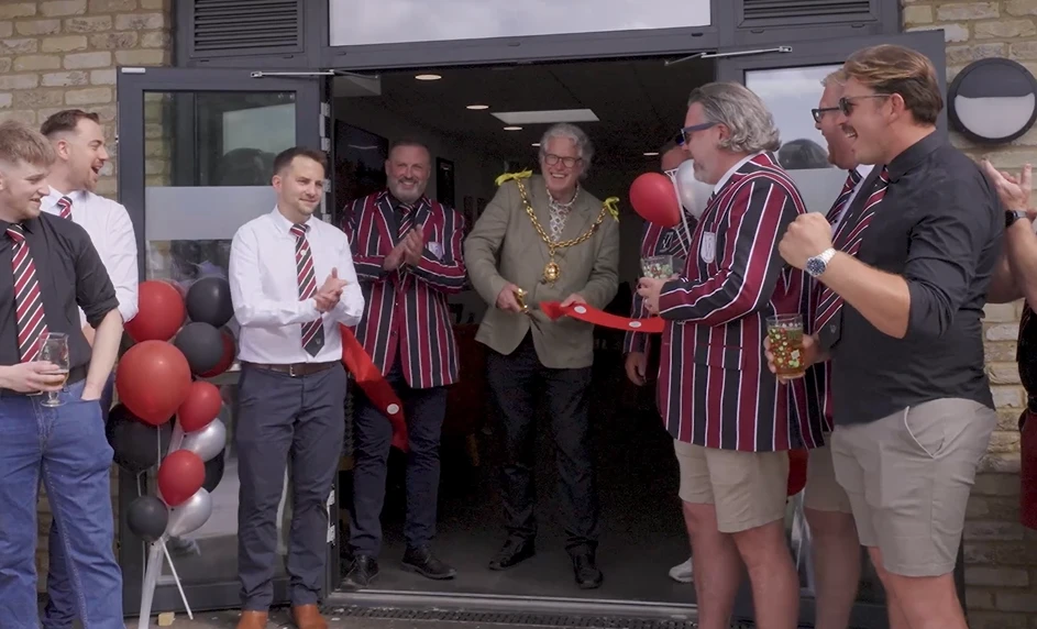 The Mayor cuts a red ribbon at the clubhouse doors, surrounded by club members in maroon and black striped blazers